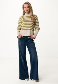 Pull rayé en crème et vert olive avec un col montant, associé à un jean large bleu foncé et des chaussures blanches à bout pointu.