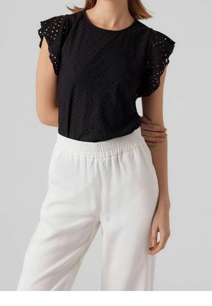 Femme portant un haut noir à manches courtes en broderie anglaise avec manches à volants et un pantalon élastique blanc taille haute sur un fond uni.