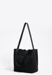 Juodas tinklelis tote maišas su dviem plačiomis rankenėlėmis, surišta viršutinė uždanga ir priekinė kišenė. Ypatybių tekstūruotas paviršius ir minimalistinis dizainas.