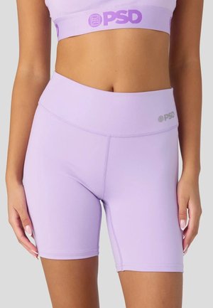 Frau trägt fliederfarbene, hoch taillierte Biker-Shorts und passenden Sport-BH mit PSD-Logos am Bund und auf der Brust vor schlichtem Hintergrund.