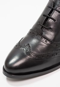 Chaussure brogue en cuir noir avec perforations décoratives et lacets noirs, vue en gros plan de l'avant et du côté.