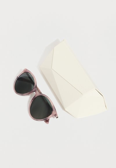 Στρογγυλά ροζ διαφανή γυαλιά ηλίου με σκούρους φακούς, τοποθετημένα δίπλα σε λευκή γεωμετρική θήκη γυαλιών ηλίου πάνω σε ανοιχτό φόντο.