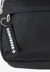 Czarny plecak z tkaniny o teksturowanej powierzchni, z metalowym zamkiem błyskawicznym i metką z napisem "CONVERSE" na białym tle. Prosty design, bez wyraźnych wzorów.