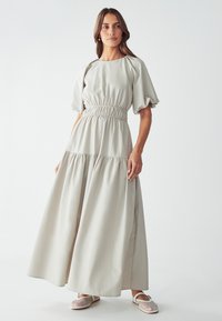 Robe maxi gris clair avec manches bouffantes, taille élastique, jupe à volants et tissu doux. Le mannequin l'a associée à des chaussures ouvertes tissées blanches.