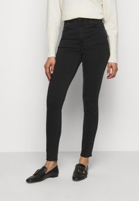 Jeans negros de talle alto con un corte ajustado, hechos de denim. Combinados con zapatos negros sin cordones que cuentan con un detalle de hebilla dorada.