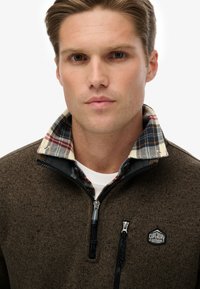 Pullover in pile marrone scuro con colletto con zip. Presenta un rivestimento in camicia a quadri nei colori blu, rosso e bianco. Include un'etichetta con logo e dettagli in metallo.