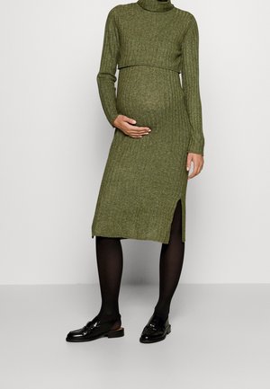 Gravid kvinde iført en grøn ribstrikket midi kjole med lange ærmer, sorte strømpebukser og sorte loafers, hånd hvilende på maven.