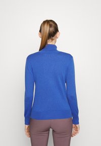 Niebieski sweter z golfem, dopasowany fason, wykonany z miękkiego materiału, z prążkowanymi mankietami i dołem, noszony z brązowymi spodniami.