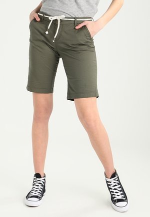 Persona che indossa pantaloncini verde oliva fino al ginocchio con cintura in corda bianca e sneakers nere alte con lacci bianchi, in piedi con le mani in tasca.