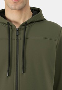 Felpa con cappuccio in tessuto verde oliva dalla trama liscia. Presenta una cerniera frontale, coulisse nere e una cucitura orizzontale sul petto.