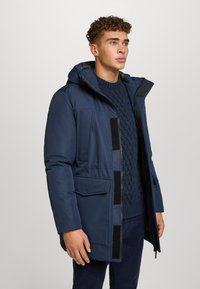 Giovane uomo che indossa un parka invernale blu navy con cappuccio sopra un maglione lavorato a maglia, in piedi su uno sfondo chiaro e uniforme.