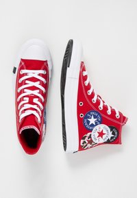 Sneakers alte in tela rossa con suole in gomma bianca, dotate di multiple toppe con il logo Converse all'esterno e lacci bianchi.