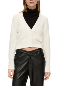 Weißer, kurzer Cardigan aus weichem, strukturiertem Stoff, mit einem tiefen V-Ausschnitt, Vorderknöpfen und einem gerippten schwarzen Rollkragenpullover darunter.