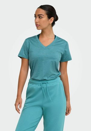 Vrouw met donker haar in een knot, die een teal V-hals T-shirt met korte mouwen en bijpassende broek met trekkoord draagt, kijkt opzij tegen een effen achtergrond.
