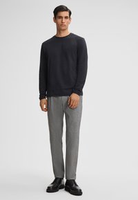 Zwarte blouse met lange mouwen, gecombineerd met lichtgrijze gestructureerde broek en zwarte veterschoenen. De blouse heeft een ronde hals en een ontspannen pasvorm.