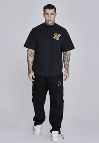 Melnas īsām piedurknēm krekls ar zelta logo uz krūtīm, saskaņots ar melnām kargo biksēm ar divām sānu kabatām un zelta akcentiem.