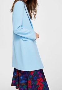 Donna che indossa un cappotto azzurro su una gonna scura con grandi motivi floreali rossi, viola e blu, mentre si trova di fronte a uno sfondo semplice.
