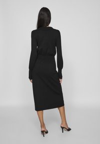 Juoda megzta suknelė su ilgomis pūstuosiomis rankovėmis, aptemptu liemeniu ir midi ilgiu. Dėvima su juodais atvirais kulnais. Lygus audinys, minimalus dizainas.