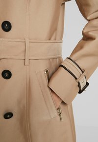 Beige trench coat featuring a belt, large black buttons, a side pocket with a gold zipper, and contrasting black accents on the sleeve.