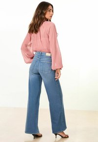 Svetle růžová blůza s texturovanými puntíky, v kombinaci s vysokým pasem a širokými modrými džínami. Outfit obsahuje přirozené tóny a hladké látky.