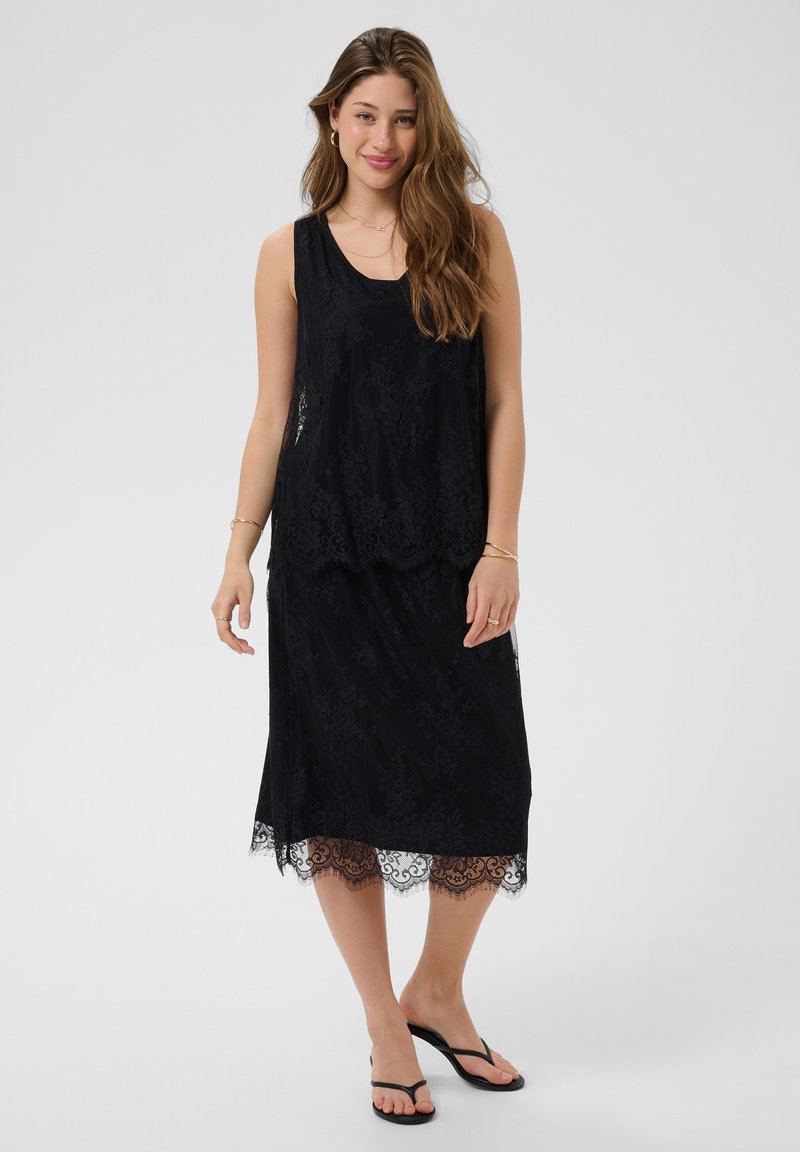 Jeune femme portant une robe noire en dentelle sans manches et des sandales noires, debout devant un fond clair uni.
