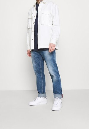 Weiße Hemdjacke mit zwei Brusttaschen, getragen über einem dunklen Hemd. Blaue Jeans mit umgeschlagenem Saum, kombiniert mit weißen Turnschuhen.
