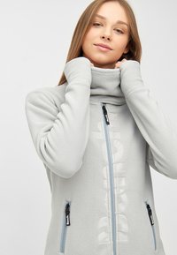 Hellgraue Fleecejacke mit hohem Kragen, durchgehendem Reißverschluss und zwei seitlichen Taschen. Verfügt über ein dezentes Logo entlang des Reißverschlusses und eine weiche Textur.