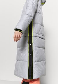 Manteau long matelassé gris avec des détails à rayures verticales noires et jaune néon, et des boutons-pression noirs, porté avec un pantalon rose.