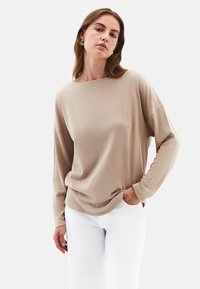 Beige langärmliges Oberteil mit lockerem Schnitt, Rundhalsausschnitt und weicher Textur, kombiniert mit weißen Hosen. Subtile Stickdetails entlang der Kanten.