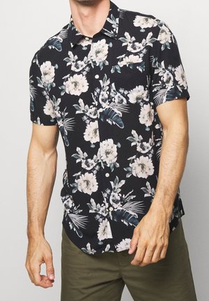 Homme portant une chemise noire à manches courtes avec de grandes fleurs blanches imprimées et un pantalon vert olive, torse visible sur un fond uni.