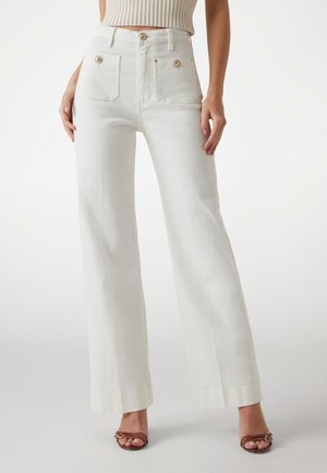 Pantaloni bianchi a vita alta e gamba larga con tasche applicate davanti e dettagli con bottoni in legno, indossati con un top corto a maglia color crema e sandali marroni con punta aperta.