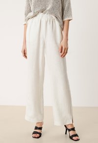 Vita linnebyxor med vida ben och elastisk midja, ihopparade med svarta klacksandaler, mot en neutral bakgrund.