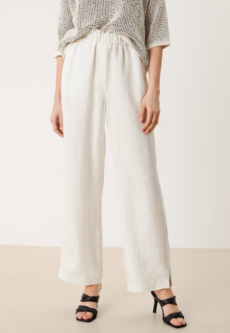 Vita linnebyxor med vida ben och elastisk midja, ihopparade med svarta klacksandaler, mot en neutral bakgrund.