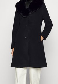 Černý kabát s umělou kožešinou na límci, přiléhavý design a dvěma předními kapsami. Má zapínání na jeden knoflík se dvěma knoflíky.