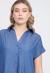 Blaues Chambray-Hemd mit Knopfleiste, kurzen hochgerollten Ärmeln und Kragen. weiche Textur mit einem subtilen Glanz.