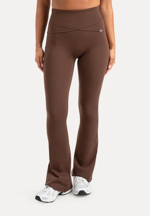Kvinde iført brune højtaljede flare leggings kombineret med hvide atletiksk sko på ensfarvet baggrund.