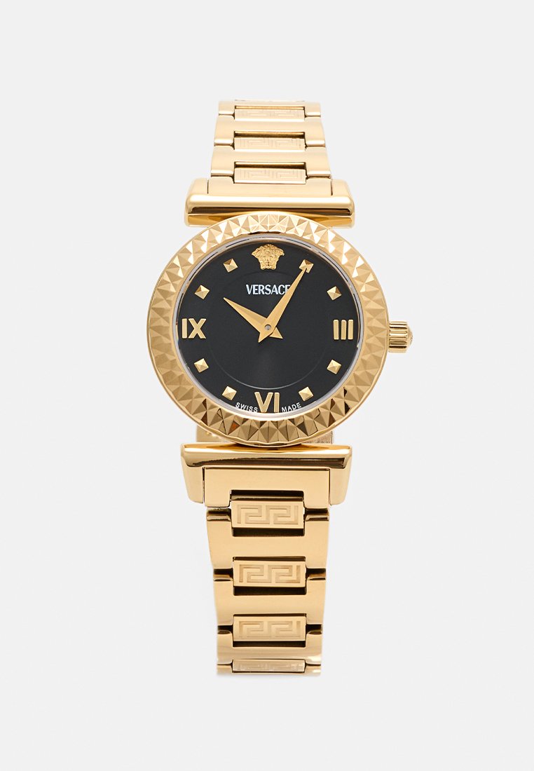 Gouden polshorloge met een zwarte wijzerplaat, Romeinse cijfers en een piramidevormige bezel. Heeft een schakelarmband met een Grieks sleutelmotief.