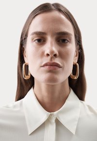 Pendientes de aro rectangulares de oro acentúan una blusa de color neutro con cuello, que presenta una textura suave y un diseño clásico con cierre de botón.