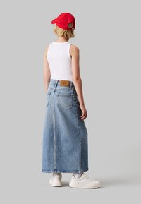 Gonna maxi in denim, azzurro, con vita alta, tasche posteriori e un logo in pelle sulla vita. Indossata con una canotta bianca e un cappellino rosso.
