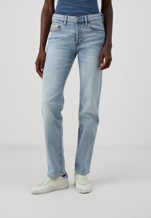 Osoba ubrana w jasnoniebieskie jeansy o prostych nogawkach i białe trampki z zielonymi sznurówkami, stojąca na jednolitym tle.