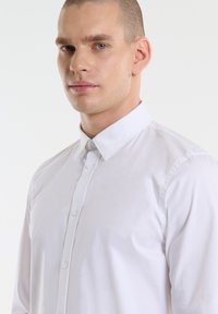 Bijela košulja s dugim rukavima i klasičnim ovratom, izrađena od glatkog materijala. Ima prednji dio s gumbima i dugim rukavima.