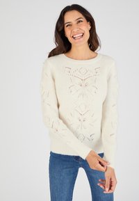 Gebreide sweater in crème, met opengewerkte bloemenpatronen, geribde mouwen en een ronde halslijn. Gecombineerd met blauwe jeans.