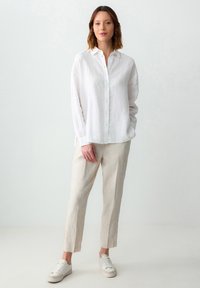 Camicia button-up bianca in lino con maniche lunghe, abbinata a pantaloni in lino beige e sneaker bianche, in piedi su uno sfondo chiaro.