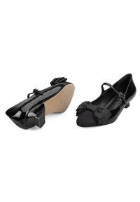 Bailarinas negras con detalles de charol y tela, puntera redondeada, tacón pequeño y lazo decorativo en la parte frontal. Incluyen una correa ajustable en el tobillo.
