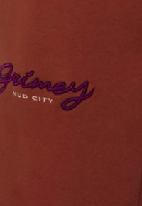 Rostfarbener Stoff mit auffällig gesticktem Text "Grimey" in Lila und "Mud City" darunter in Weiß, geschwungener Schriftart.