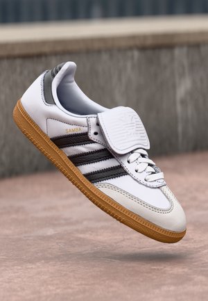 Valkoinen Adidas Samba -lenkkari mustilla raidoilla, kumipohjalla ja suurella kielekkeellä, leijailee betonipinnan yläpuolella matalan seinän vieressä.