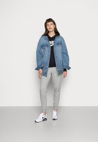 Giacca di jeans blu chiaro, vestibilità oversized con tasche; felpa nera sotto con logo bianco; pantaloni della tuta grigi; sneakers bianche con dettagli blu navy.