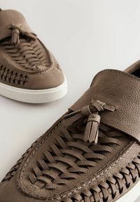 Brown leather slip-on shoes with woven design, tassel detail, and white rubber sole. Textured surface with contrasting stitching.