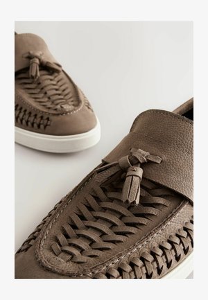 Scarpe slip-on in pelle marrone con design intrecciato, dettaglio con nappina e suola in gomma bianca. Superficie testurizzata con cuciture a contrasto.