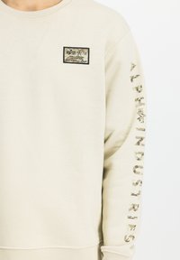 Béžová mikina vyrobená z jemnej tkaniny, s kamuflážnou nášivkou na hrudi a s potlačou "ALPHA INDUSTRIES" v kamuflážnom dizajne na rukáve.